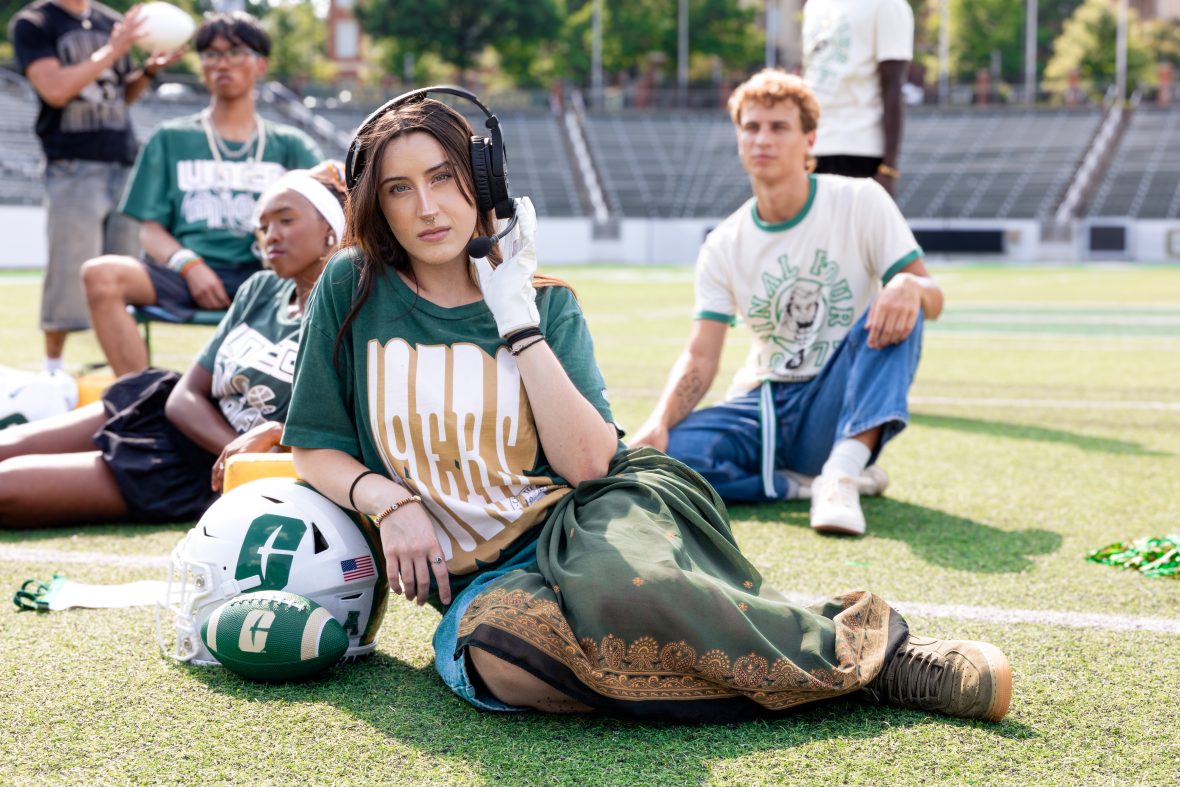 Student sitting on ground with their legs to the side, wearing charlotte apparel and resting on various sports balls